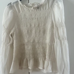 Vit blus från Vero Moda - Säljer en söt vit blus från Vero Moda med smockdetaljer och volangkant. Blusen har långa ärmar och en luftig passform, perfekt för både vardag och fest. Den är tillverkad i ett lätt och bekvämt material.