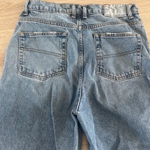Blå jeansbyxor - Säljer ett par klassiska blå jeansbyxor med femficksdesign och bälteshällor. Byxorna har en rak passform och är perfekta för en avslappnad stil. De är tillverkade i slitstarkt denim och har en traditionell dragkedja och knapp framtill.