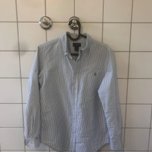 Vit och blå randig skjorta från Ralph Lauren - Klassisk vit och blå randig skjorta från Ralph Lauren med knappar framtill och en broderad logotyp på bröstet. Skjortan har långa ärmar och är tillverkad i ett bekvämt material. Bra skick 