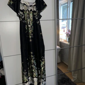 Svart långklänning med blommönster - Elegant svart långklänning från Vali Tricot med vackert blommönster i grönt och beige. Klänningen har korta ärmar och en knytning framtill. Perfekt för en stilfull look.