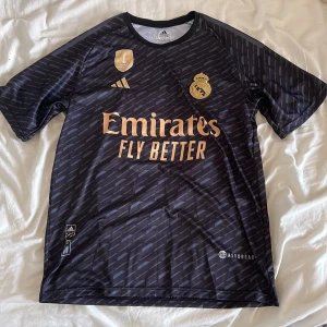 Svart Real Madrid fotbollströja - Snygg svart Real Madrid fotbollströja från Adidas med guldiga detaljer och text. Tröjan har korta ärmar och ett diskret mönster. På framsidan finns klubbens emblem och sponsorlogga, medan baksidan pryds av 'VINI JR.' och nummer 7.