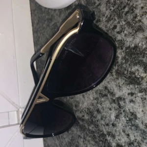 Svarta solglasögon från Louis Vuitton - Snygga svarta solglasögon från Louis Vuitton med gulddetaljer. De har en modern design med en tjock ram och mörka glas. Perfekta för att ge en lyxig touch till din stil.