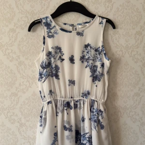 Vit blommig klänning från KappAhl - Säljer en söt vit klänning från KappAhl med blått blommönster. Klänningen är ärmlös och har en luftig passform med en liten knapp i nacken. Perfekt för varma dagar! 🌸