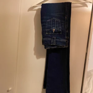 Mörkblå jeans från Gina Tricot - Mörkblå jeans från Gina Tricot med kontrasterande sömmar och fickor med knappdetaljer. Perfekta för en stilren look. De har en klassisk design med bälteshällor och knappgylf.