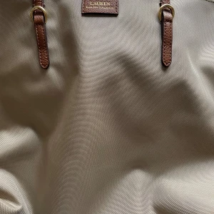 Beige shoppingväska från Ralph Lauren - Säljer en stilren beige shoppingväska från Ralph Lauren. Väskan har bruna skinnhandtag och en praktisk dragkedja i guld. Perfekt för en dag på stan eller som en elegant vardagsväska.