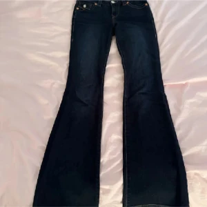 Mörkblå bootcut jeans från True Religion - Snygga mörkblå bootcut jeans från True Religion med klassisk femficksdesign och märkets ikoniska broderade bakfickor. Jeansen har en låg midja och är perfekta för en avslappnad stil. De har orange kontrastsömmar och en patch med logotyp baktill.