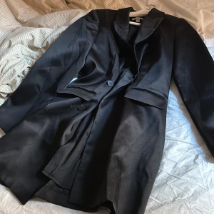 Svart kavaj-klänning  från H&M - Snygg svart kavaj från H&M i storlek S. Kavaj- klänningen  har en klassisk design med knappar framtill och fickor. Perfekt för en stilren look.