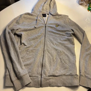 Grå hoodie från Lyle & Scott storlek S - Säljer en stilren grå hoodie från Lyle & Scott med dragkedja och huva. Tröjan har ett broderat emblem på bröstet och är tillverkad i mjuk bomull. Perfekt för en avslappnad stil.