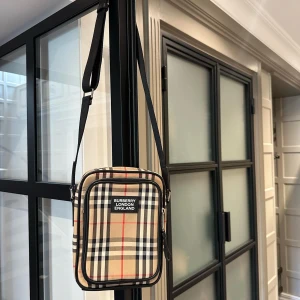 Vintage Burberry väska - Säljer en snygg rutig axelväska från Burberry med klassiskt mönster i beige, svart och rött. Väskan har en praktisk dragkedja och justerbar axelrem. 