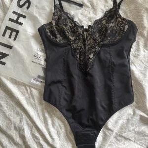 Shapewear body - Supersnygg svart shapewear body från Shein med djup v-ringning framtill i fin spets. Snygga sömdetaljer och hakknäppning nedtill. Perfekt för att lyfta vilken outfit som helst. Helt ny och aldrig använd då jag köpte fel storlek.