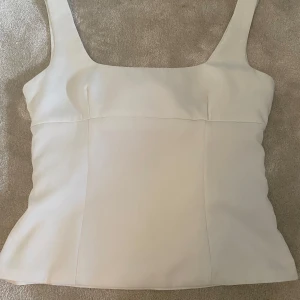 Vit linnetopp med rosett i ryggen - Supersnygg vit linnetopp med breda axelband och fyrkantig ringning. Baksidan har en stor rosett och en öppen detalj, plus en diskret dragkedja i sidan. Toppen är gjord i ett slätt, lätt glansigt material som känns lyxigt och passar perfekt till jeans eller kjol. Lite lös i midjan på mig.