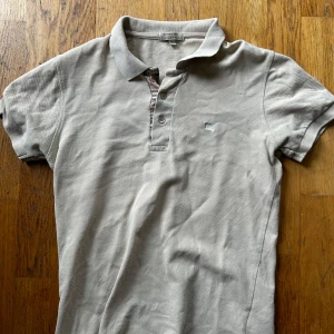 Beige Burberry slim fit piké XS - Snygg beige pikétröja från Burberry i slim fit-modell. Klassisk krage, korta ärmar och knappar framtill med den ikoniska Burberry-rutiga insidan. Liten broderad logga på bröstet. Tillverkad i 100% bomull. 