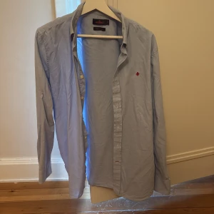 Ljusblå skjorta från Morris, M - Snygg ljusblå skjorta från Morris med klassisk krage och knappar framtill. Broderad röd detalj på bröstet och tillverkad i bomull. Perfekt för dig som gillar stilrena plagg med en twist.