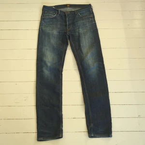 Lee Daren Button Fly jeans blå W31 L34 - Snygga blå jeans från Lee, modell Daren Button Fly, med klassisk femficksdesign och Lee-logga på bakfickan. Jeansen har normal passform, raka ben och är tillverkade av ekologisk bomull. Färgen är mörkblå med snygga slitningar.