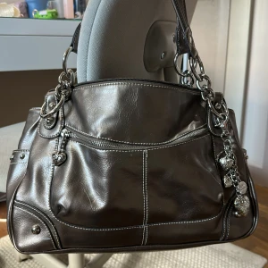 Brun axelväska med hjärtberlocker - Snygg brun axelväska i skinn med silvriga detaljer och dekorativa hjärtberlocker. Väskan har dragkedja framtill, markerade sömmar och nitar på sidorna. Perfekt storlek för vardagens essentials och ger en cool vibe med sina metallringar och kedjor. Får platts med dator! 🩷🩷