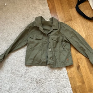 Olivgrön overshirt från Ü, stl 146 - Olivgrön overshirt från Ü med klassisk krage, två stora bröstfickor med knappar och raka ärmar. Jackan har en avslappnad passform och stängs med knappar framtill. Perfekt för lager-på-lager och enkel att styla till olika outfits.