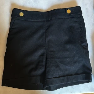 Svarta shorts från H&M med guldknappar - Svarta shorts från H&M i storlek 34 med snygga guldknappar framtill. De har hög midja, uppvikta benslut och diskreta bakfickor. 