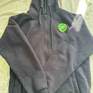 Hammarby hoodie - Svart hoodie med dragkedja och huva, Hammarby-emblem på bröstet och stort tryck på ryggen med texten 'HAMMARBY' och illustration av personer i hoodie. Perfekt för dig som vill visa ditt stöd för laget. Mjuk insida och ribbade muddar.