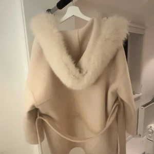Beige kappa med pälshuva - Säljer en elegant beige kappa med en lyxig pälshuva. Den har långa ärmar och ett bälte i midjan för en snygg passform. Perfekt för kyliga dagar när du vill hålla stilen. Har några fläckar på sig som jag tror hade gått bort i tvätten. Passar även M/L