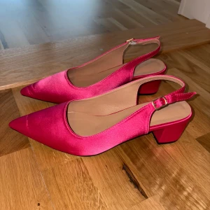 Rosa pumps med spetsig tå och klack - Snygga pumps i starkt rosa satin med spetsig tå och låg klack. Skorna har öppen häl med justerbart spänne och en elegant, stilren design som verkligen sticker ut. Perfekta för dig som vill addera färg till din outfit. Storlek 42 men sitter som 40