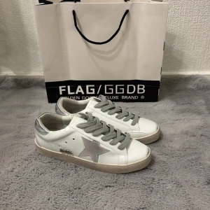 Golden Goose Superstar sneakers vit/grå - Golden Goose Superstar sneakers i vitt skinn med grå mockadetaljer och silverfärgad häl. Grå snörning och ikonisk stjärna på sidan. Platt sula och rund tå ger en clean och cool look. Perfekta för dig som gillar streetstyle med lyxig känsla.
