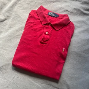 Röd pikétröja från Polo Ralph Lauren - Säljer en klassisk röd pikétröja från Polo Ralph Lauren i storlek L. Tröjan har krage, knäppning med två knappar och den ikoniska broderade loggan på bröstet. Tillverkad i mjuk bomull som är skön mot huden. Perfekt för en stilren och avslappnad look.