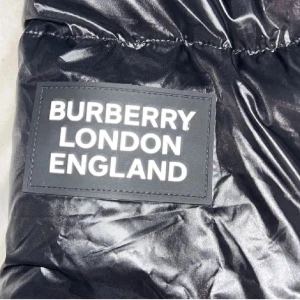 Svart pufferjacka från Burberry - Säljer en svart pufferjacka från Burberry med glansig finish och klassiskt rutigt foder i svart, vitt och rött. Jackan har dragkedja framtill och fickor med dragkedja. Perfekt för dig som vill ha en snygg och varm jacka med ikonisk Burberry-stil.