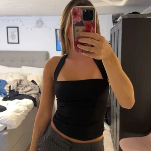 Svart halterneck topp - Snygg svart halterneck topp med breda axelband och öppen rygg. Toppen är tight och figurnära, perfekt att styla med både jeans och kjol. Enkel och stilren design som passar till många olika looks. Använd 2 gånger