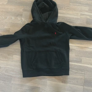 Svart hoodie från Polo Ralph Lauren - Svart hoodie från Polo Ralph Lauren med klassisk röd broderad logga på bröstet. Tröjan har huva, känguruficka och långa ärmar. Mjuk och skön bomullskvalitet, perfekt för chill dagar. Snygg och enkel design som funkar till det mesta.