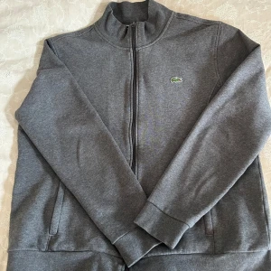 Grå ziptröja från Lacoste Sport XL - Snygg grå ziptröja från Lacoste Sport i storlek XL. Tröjan har hel dragkedja, ståkrage och det klassiska gröna krokodilmärket på bröstet. Perfekt för en chill och sportig stil. Mjuk och skön bomullsblandning med långa ärmar och fickor på sidorna.
