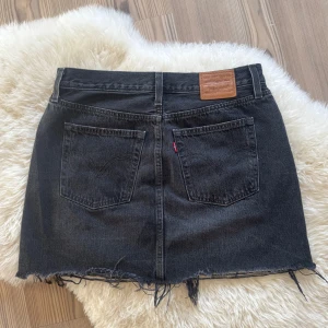 Svart jeanskjol från Levi's - Cool svart jeanskjol från Levi's med rå, fransig kant och klassiska fem fickor. Kjolen har bälteshällor, knapp och dragkedja framtill samt Levi's ikoniska läderpatch bak. Perfekt för en avslappnad och trendig look.