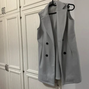 Ljusgrå lång väst med knappar - Snygg ljusgrå väst med klassisk krage och svarta knappar framtill. Västen är lång och har en stilren, rak passform som ger en cool look. Perfekt att slänga över en hoodie eller tröja för extra lager. Materialet känns mjukt och har en lite dressad vibe.