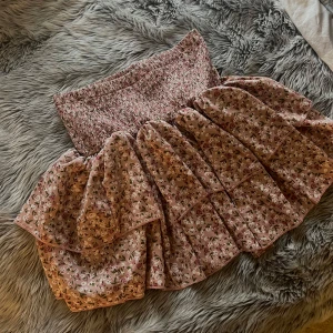 Rosa blommig volangkjol - Supersöt kort kjol i ljusrosa med små blommor och volanger. Kjolen har en smockad midja som sitter bekvämt och ger en snygg passform. Perfekt för dig som gillar romantisk och feminin stil.