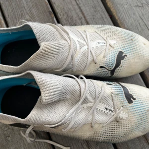 Vita Puma Future fotbollsskor - Säljer ett par vita Puma Future fotbollsskor med stickad ovandel och snörning. Skorna har svarta Puma-loggor, detaljer i ljusblått och en sula med rosa dobbar. Perfekta för dig som vill ha lätta och smidiga skor på planen.