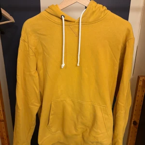 Gul hoodie med huva och ficka - Gul hoodie med klassisk känguruficka och vit snörning i huvan. Tröjan har lång ärm och en avslappnad passform, perfekt för chill dagar. Tillverkad i mjukt bomullsmaterial som känns skönt mot huden. Storlek M