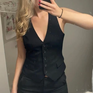 Svart kostymväst från H&M - Stilren svart väst från H&M med djup v-ringning och knappar framtill. Klassisk kostymlook med två fickor och figurnära passform. Perfekt att styla över en skjorta eller t-shirt för en trendig vibe.