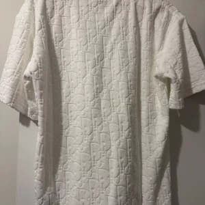 Vit Dior t-shirt i frotté - Exklusiv vit t-shirt från Dior i mjuk frotté med diskret mönstrad logga över hela plagget. T-shirten har korta ärmar och en relaxed fit som ger en chill vibe. Perfekt för dig som vill sticka ut med en lyxig och unik look. aldrig använd 