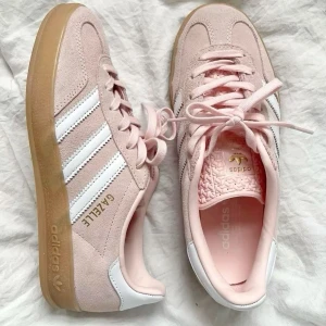Adidas Gazelle rosa mockasneakers - Snygga Adidas Gazelle sneakers i ljusrosa mocka med klassiska vita ränder på sidorna och gummisula. Skorna har snörning och en retrovibe som passar perfekt till streetwear. Guldiga detaljer och mjuk insida för extra komfort.