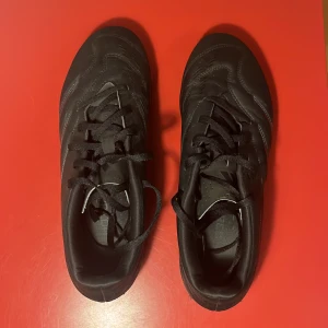Svarta adidas fotbollsskor 43 1/3 - Snygga svarta fotbollsskor från adidas i storlek 43 1/3. Skorna har klassisk snörning, mönstrad ovandel i syntet och fasta dobbar för bra grepp på planen. Perfekta för dig som vill ha en clean look på matchen eller träningen.