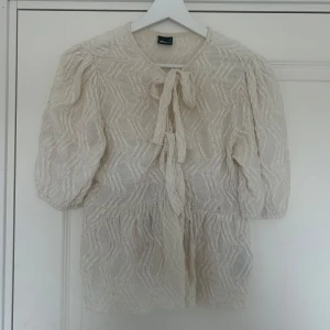 Beige blus med knyt och puffärm - Supersöt beige blus från Gina Tricot med strukturmönster, puffiga halvlånga ärmar och två knyt framtill. Blusen har en lätt volang nedtill och är gjord i ett tunt, luftigt material som känns perfekt för vår och sommar.