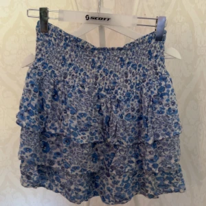 Blommig blå kjol från BikBok - Supersöt kjol från BikBok med blått och vitt blommönster. Kjolen har volanger och smock upptill för en bekväm passform. Perfekt för varma dagar när du vill känna dig både chill och stylish.