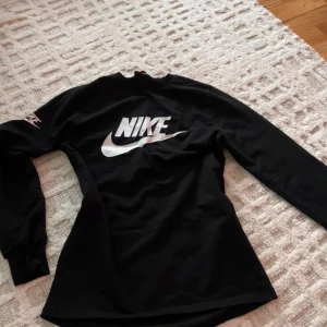 Svart långärmad Nike tröja - Svart långärmad tröja från Nike med stor vit logga på bröstet och mindre logga på ärmen. Tröjan har en klassisk passform och är gjord i mjuk bomull, perfekt för en sportig och avslappnad stil.