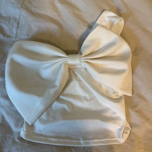 Vit axelbandslös topp med stor rosett - Supersöt vit topp med en stor rosett framtill och one shoulder-design. Perfekt för dig som vill sticka ut med en unik och trendig look. Toppens material känns mjukt och har en lätt glans. Snygg att matcha med både jeans och kjol.