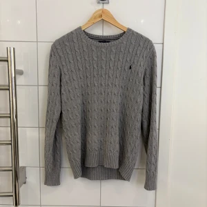 Grå stickad tröja från Polo Ralph Lauren - Säljer en stilren grå stickad tröja från Polo Ralph Lauren. Tröjan har ett klassiskt flätmönster och är långärmad med ribbade muddar. Perfekt för kyligare dagar.