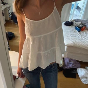 Vit volangig linnetopp med tunna band - Supersöt vit linnetopp med tunna axelband och volangiga lager. Toppen är luftig och har en flowy passform, perfekt för varma dagar. Materialet känns lätt och svalt mot huden. Enkel att matcha med jeans eller shorts för en chill look.