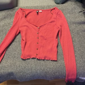 Rosa ribbad topp från H&M Divided - Säljer en rosa ribbad topp från H&M Divided med croppad passform och v-ringning. Toppen har långa ärmar, knappar framtill och vågig kant nertill. Perfekt för dig som gillar en enkel men ändå trendig stil.
