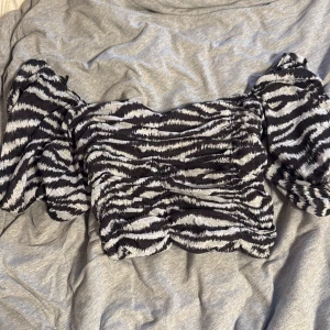 Zebramönstrad topp från Pull&Bear - Säljer en svartvit zebramönstrad topp från Pull&Bear med puffiga korta ärmar och smockad, croppad passform. Toppen har vågig nederkant och är perfekt för dig som gillar att sticka ut med djurmönster. Superstretchig och bekväm att bära.