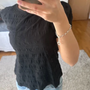 Svart blus med puffärm och struktur - Svart blus med korta puffärmar och snygg struktur i tyget. Modellen har rund halsringning och en liten knapp i nacken. Tyget är lätt och har en vågig, texturerad känsla som ger en cool look till outfiten.