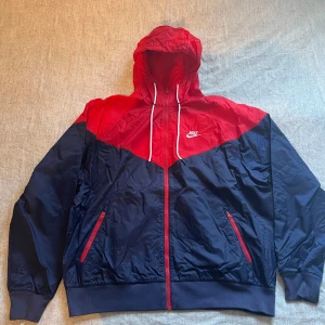Nike vindjacka - Nike vindjacka med huva i rött och marinblått, perfekt för blåsiga dagar. Säljer jackan för att jag är inte i behov av den längre. Vid snabb affär kan priset diskuteras!