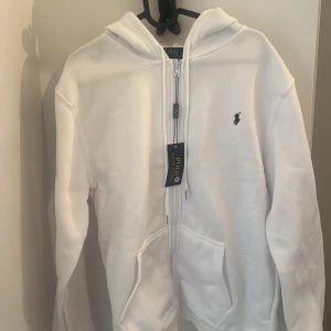 Vit hoodie från Polo Ralph Lauren - Snygg vit hoodie från Polo Ralph Lauren med dragkedja och klassisk blå logga på bröstet. Tröjan har huva, långa ärmar och två stora fickor framtill. Perfekt för en chill och stilren look.